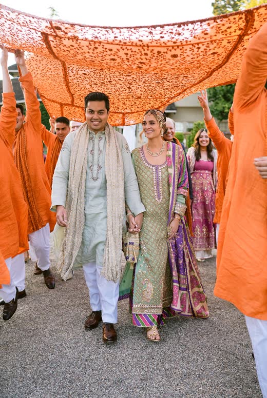 The couple arriving under an orange canopy carried by guests